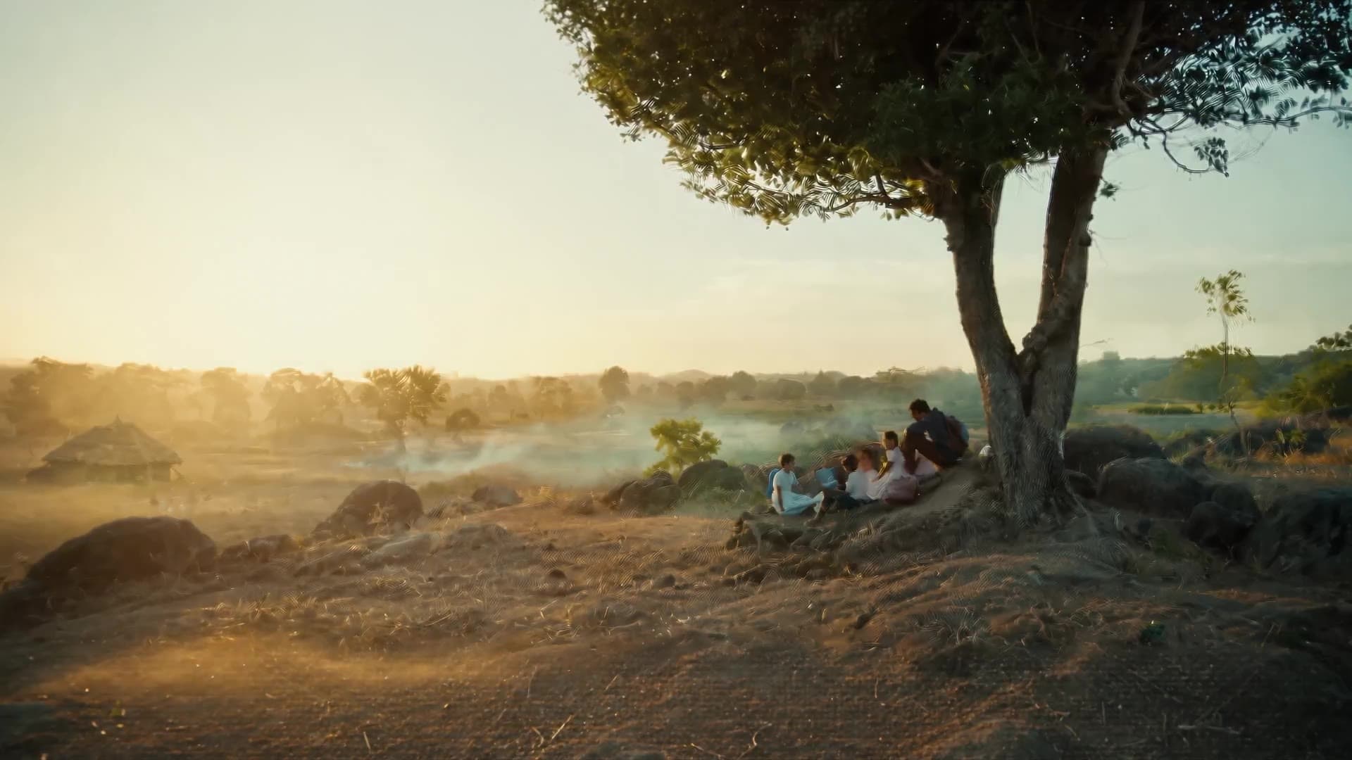 Schoolchildren sitting under a tree at first light, mist drifting over a village and open country behind them.